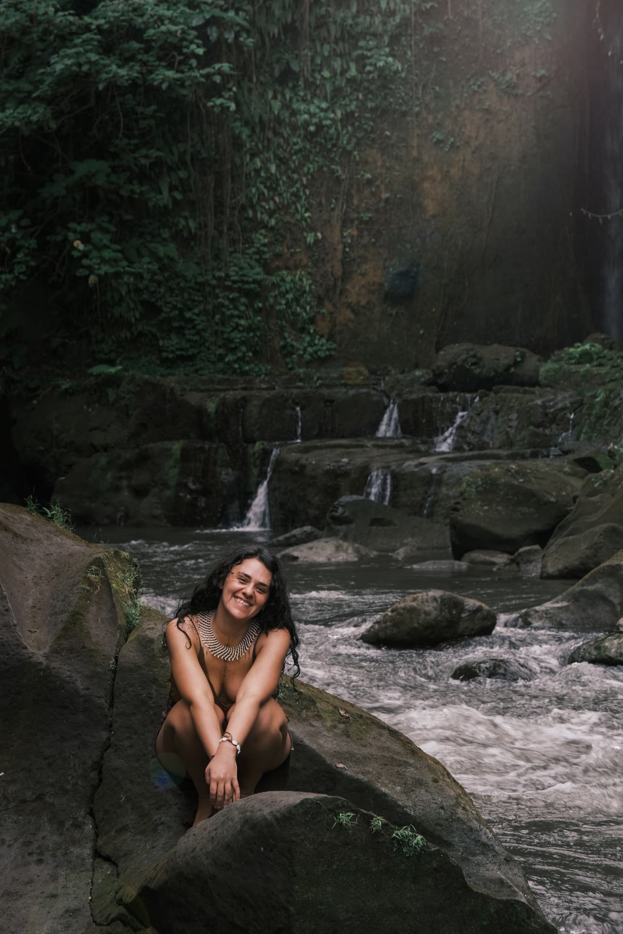 Nicole seated on mossy rocks by a waterfall in the forest, smiling